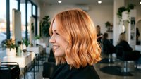 Happy blonde woman with wavy bob haircut smiling in modern hair salon with stylists working in background. Professional beauty and haircare service concept.