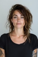 Young woman with tousled brown hair and tattoos wearing black t-shirt against neutral background, natural makeup look for fashion or lifestyle content.