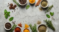 Aromatic spices and fresh herbs arranged on textured stone surface. Culinary ingredients including turmeric, paprika, peppercorns, cinnamon, rosemary, thyme.