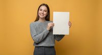 Young Caucasian woman in gray sweater holding blank white paper sheet against orange background, smiling at camera for mockup presentation design.