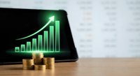 Digital tablet displaying glowing green growth chart with upward arrow and stacked golden coins on wooden desk representing financial success and investment profits.