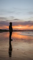 Silhouetted male figure standing on wet sand beach at sunset, gazing toward ocean horizon with dramatic golden and purple sky, contemplative mood, suitable for editorial and lifestyle concepts.