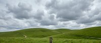 Rolling green hills under dramatic cloudy sky with wooden fence post in foreground, suitable for landscape and nature backgrounds.