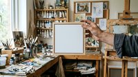 Senior male artist holding blank canvas in creative studio workspace with art supplies, paintbrushes, and wooden furniture for creative projects and artistic endeavors.