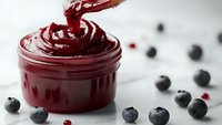 Rich blueberry jam being poured into glass jar with fresh berries scattered around on white marble surface for gourmet cooking and breakfast recipes.