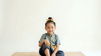 Young Asian girl, approximately 5-7 years old, wearing casual denim shirt and jeans, sitting on wooden table against white background, smiling at camera with cheerful expression, suitable for children's education, family lifestyle, and parenting content.