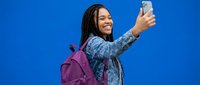 Young African American woman with braids taking selfie with smartphone against bright blue background wearing denim jacket and purple backpack for social media content.