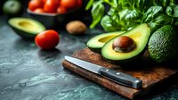 Fresh avocado halves on wooden cutting board with knife, surrounded by organic vegetables including tomatoes and leafy greens for healthy cooking preparation.