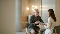 Mature man with gray hair and light skin tone, wearing olive blazer, engaged in conversation with young woman with long dark hair and light skin tone wearing cream blouse in modern office setting with warm lighting. Business meeting or professional discussion.