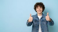 Happy teenage boy with curly hair showing thumbs up gesture in denim jacket on blue background for positive youth marketing campaigns.