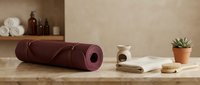 Burgundy yoga mat rolled on wooden surface with wellness accessories including towel, brush and plant in minimalist spa setting for relaxation practice.