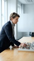 Mature female professional in navy blazer examining architectural model on wooden table in modern office with bright windows, representing real estate development and urban planning.