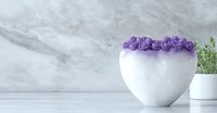 White heart-shaped vase adorned with purple roses on marble counter, minimalist home decor with soft green plant accent creates serene romantic atmosphere.