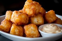 Golden crispy potato tots piled on white plate with creamy dipping sauce, close-up view showing crunchy exterior and fluffy interior against dark background.