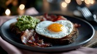 Rustic breakfast with sunny side up egg, crispy bacon, and fresh avocado on dark toast served on black plate with soft bokeh lights in background.