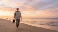 Middle-aged Caucasian male professional walking barefoot on sandy beach at sunset, carrying shoes and briefcase, representing work-life balance and career transition concepts.