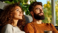 Young diverse couple relaxing at home near window in warm sunlight. Hispanic woman with curly hair and bearded man in brown sweater enjoying peaceful moment.