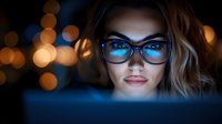 Young woman with glasses working at night on computer with blue screen reflection and bokeh lights in background.