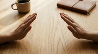 Two hands positioned face to face in prayer or meditation gesture on wooden desk with coffee cup and leather notebook creating peaceful workspace atmosphere.