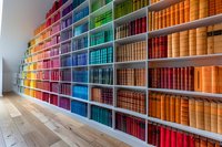 Colorful rainbow bookshelf with books arranged by spectrum colors from purple to red, creating vibrant library display on wooden floor.