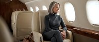 Professional blonde businesswoman in elegant gray suit relaxing in luxury private jet cabin during flight for corporate travel and business aviation.