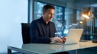 Middle-aged Caucasian businessman working late on laptop in modern office with city view. Professional executive analyzing data in corporate workspace.