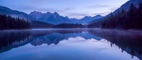 Serene mountain lake at dawn with perfect reflections of snow-capped peaks and forest silhouettes in calm blue waters creating peaceful alpine landscape.