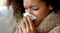Young African American woman with curly hair blowing nose in tissue while wrapped in cozy blanket, showing cold and flu symptoms during illness at home.