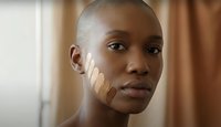 Young African American woman applying foundation makeup with fingers on face in warm indoor lighting for beauty and skincare routine.
