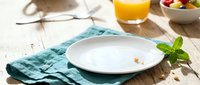 Empty white plate with teal napkin on wooden table, fresh mint garnish, orange juice and fruit bowl nearby, breakfast or lunch setting.
