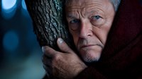 Senior Caucasian man with grey hair and wrinkled face wearing red cloak embraces tree trunk in dramatic moody lighting with blue bokeh background, expressing connection to nature.
