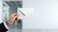 Male hand in business suit holding white envelope against blurred office background, representing mail delivery, correspondence, communication, and professional messaging for corporate concepts.