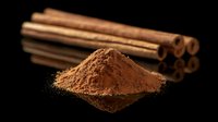 Ground cinnamon powder heap and whole aromatic spice sticks on black reflective surface with dramatic lighting, macro food photography.