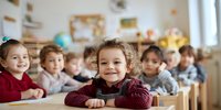 Diverse group of preschool children sitting at desks in classroom, focus on smiling young child in burgundy sweater in foreground.