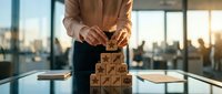 Businesswoman stacking wooden blocks with icons into a pyramid on a glass desk in a modern office, symbolizing strategy and growth.