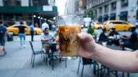 Caucasian male hand holding cold cola beverage with ice in urban street setting, busy city environment with yellow taxis and pedestrians in background, refreshment concept for advertising and lifestyle use.