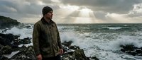 Man in olive jacket standing on rocky coastline with dramatic stormy ocean waves and cloudy sky, suitable for travel, lifestyle, and outdoor adventure content.