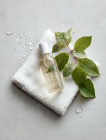 Natural cosmetic oil bottle with fresh green leaves on white towel against light background with water drops, spa and wellness concept.