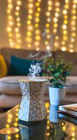 Decorative white aroma diffuser with geometric pattern emitting steam, set against warm fairy lights background with houseplant and cozy living room decor.