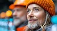 Mature woman with gray beard wearing orange knit beanie smiling and looking up thoughtfully against blurred urban background with bokeh lights. Concept of gender identity.