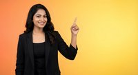 Professional South Asian businesswoman in black suit pointing upward on vibrant orange yellow gradient background for corporate presentations.