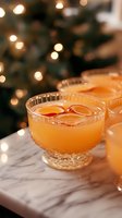 Orange citrus cocktail in crystal coupe glass garnished with fresh citrus slices on marble surface, with festive bokeh lights and Christmas tree in background.