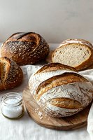 Fresh artisanal bread loaves with crispy crust on wooden board, sliced sourdough, sesame seed topped buns and sea salt in glass jar, rustic food styling.