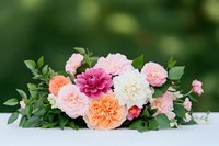 Fresh floral arrangement featuring pink and peach garden roses, burgundy dahlia, and white peony with green foliage on light surface against blurred natural background.