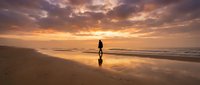 Silhouette of a woman standing on wet sand at sunset, gazing at the ocean horizon during golden hour. Suitable for wellness, travel, and lifestyle content.