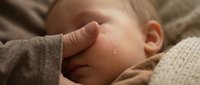 Newborn baby crying with a teardrop on cheek, adult hand gently touching face, soft warm tones, emotional parenting moment.
