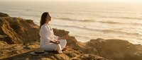 Woman meditating in lotus pose on coastal cliff at golden hour sunset, peaceful mindfulness practice by ocean waves for wellness and spiritual balance.