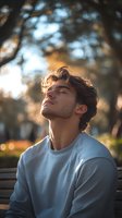 Young man with closed eyes enjoying peaceful moment in park during golden sunset, wearing casual white sweatshirt, with wind-swept curly hair.