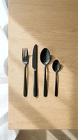 Black matte flatware set arranged on light wood surface with natural sunlight casting shadows. Four-piece cutlery collection featuring fork, knife, and two spoons for dining and tableware design.