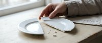 Hand sweeping crumbs from white plate into napkin on wooden table by window. Kitchen cleanup and table maintenance after meal.